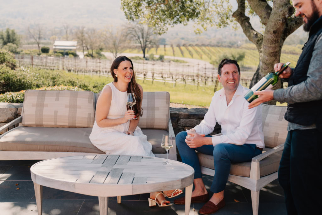 A seated man and woman, both dressed in white and seated outside, smile as a man presents a bottle.